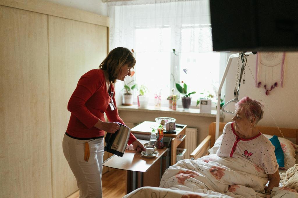 Ambiente doméstico; quarto bem iluminado de idosa convalescente, que está na cama; cuidadora sorridente serve café matinal.